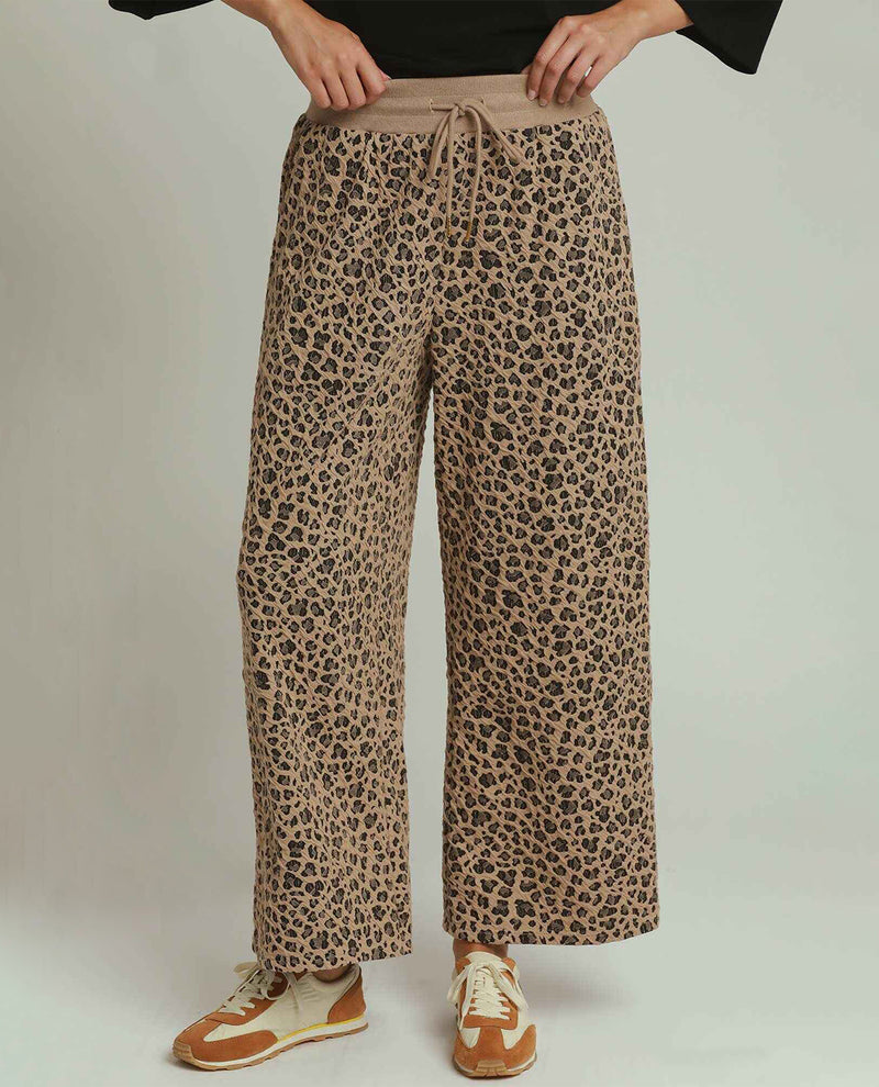 A person wearing wide-leg leopard-print pants with a drawstring waistband, paired with brown and cream sneakers, shown from the waist down against a plain light gray studio background.