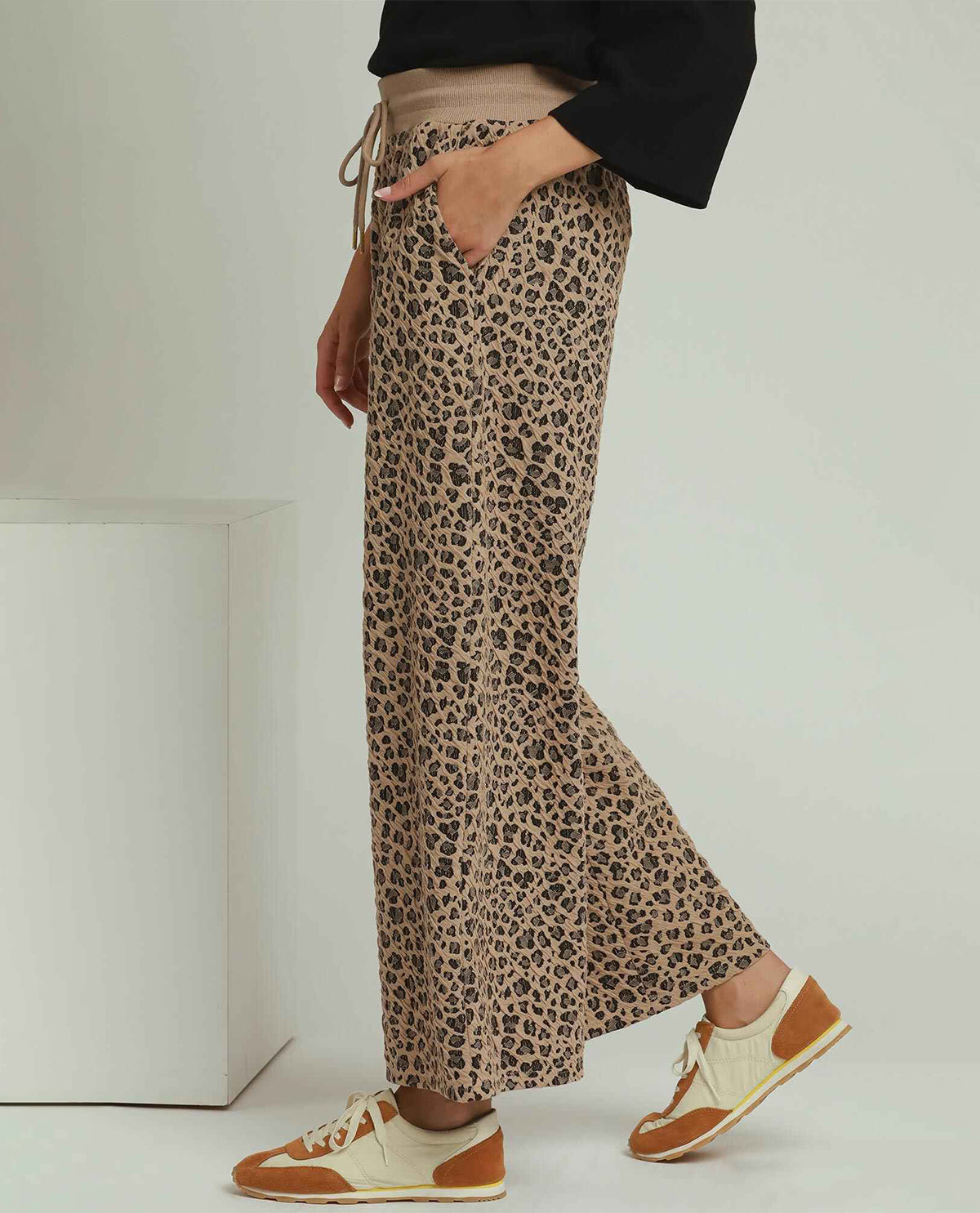 A person wearing loose, wide-leg leopard-print pants with a beige drawstring waist, paired with a black top and tan-and-cream sneakers, photographed in a clean studio setting.