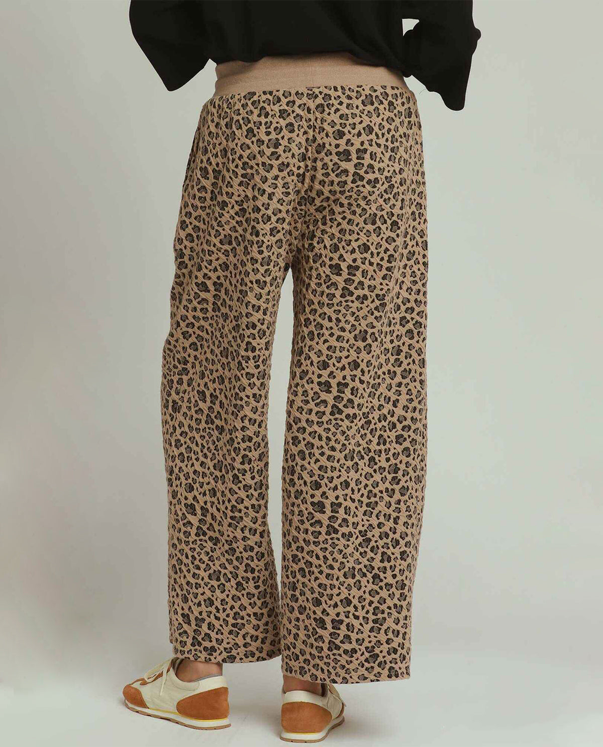 A person shown from the back wearing wide-leg leopard-print pants, a black top, and white and tan loafers against a plain light gray studio background.
