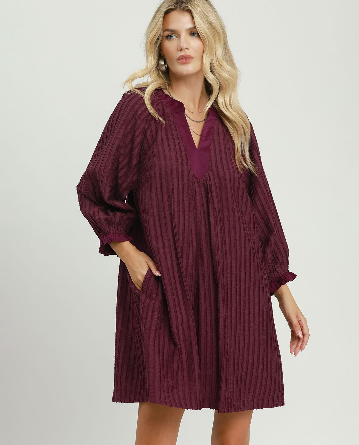 A woman modeling a loose-fitting, long-sleeve burgundy tunic dress with vertical textured stripes, a split neckline, and side pockets against a light gray studio background.