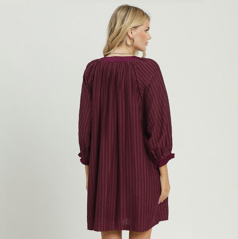 A woman shown from the back wearing a pleated, long-sleeve dress in a deep burgundy or maroon color, set against a plain light gray studio background.