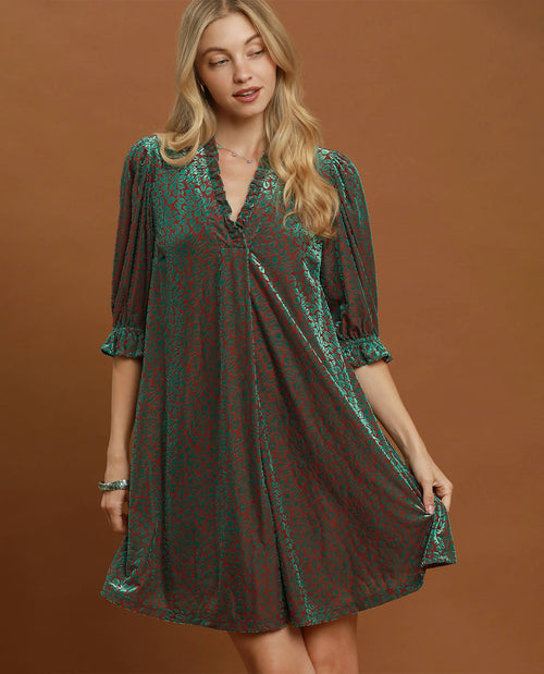 A woman modeling a short, loose-fitting dress with three-quarter sleeves and a V-neckline, featuring a green and red abstract pattern, posed against a solid brown studio backdrop.