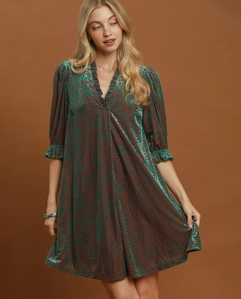 A woman modeling a short, loose-fitting dress with three-quarter sleeves and a V-neckline, featuring a green and red abstract pattern, posed against a solid brown studio backdrop.