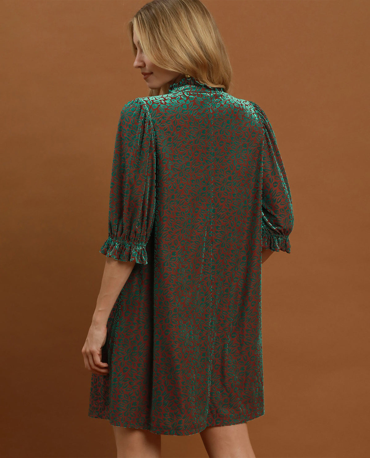 A woman wearing a short-sleeve dress with a green and red abstract pattern, shown from the back against a solid brown studio background.