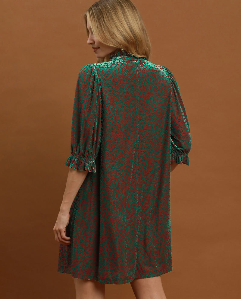 A woman wearing a short-sleeve dress with a green and red abstract pattern, shown from the back against a solid brown studio background.