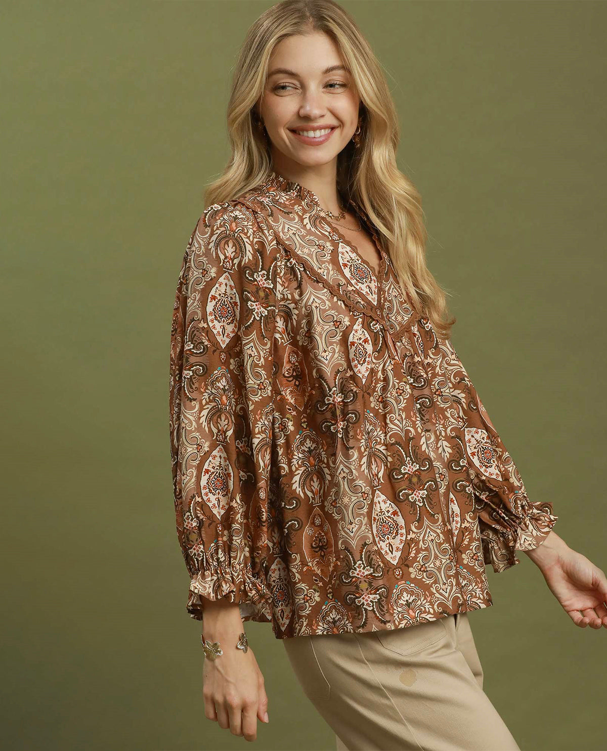 A woman modeling a brown paisley pattern blouse with beige and white details, paired with beige pants, against a solid olive green background.