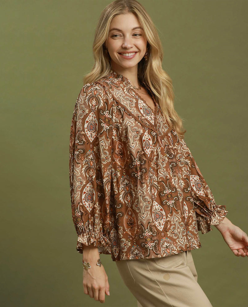 A woman modeling a brown paisley pattern blouse with beige and white details, paired with beige pants, against a solid olive green background.