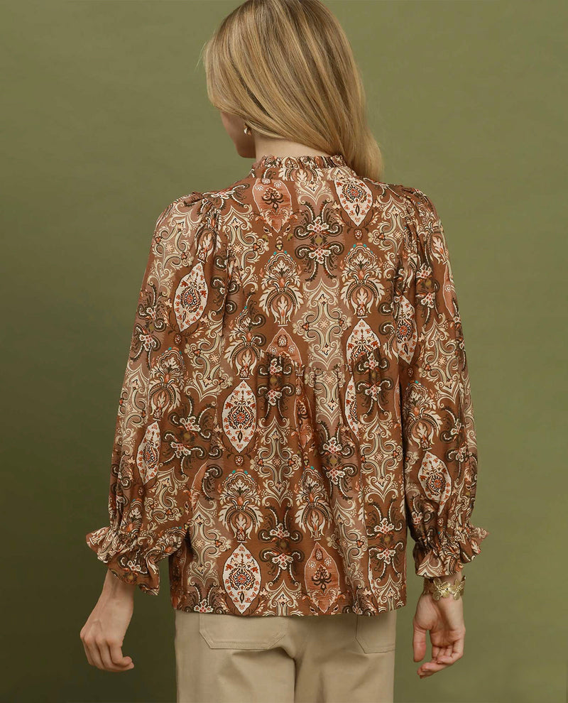 A woman viewed from the back wearing a brown and beige patterned blouse with long ruffled sleeves, paired with beige pants, against an olive green background.