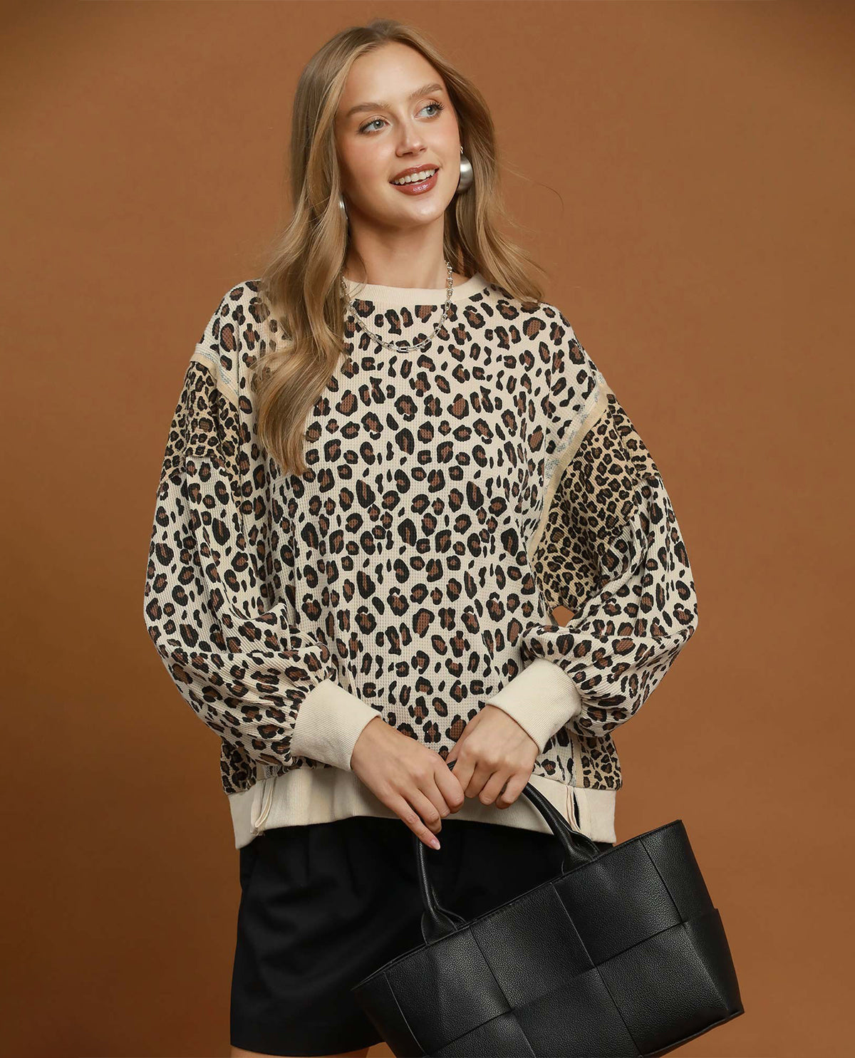 A woman modeling a leopard print and solid beige knit sweater with black shorts and a black woven tote bag, set against a solid brown studio background.