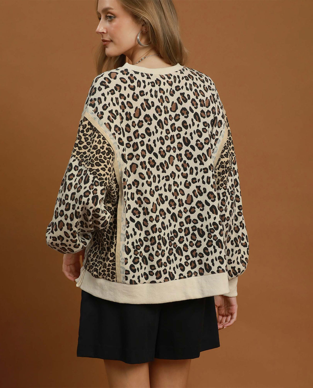 A woman shown from the back wearing a leopard-print sweatshirt with beige side panels and black shorts, posed against a solid brown studio background.