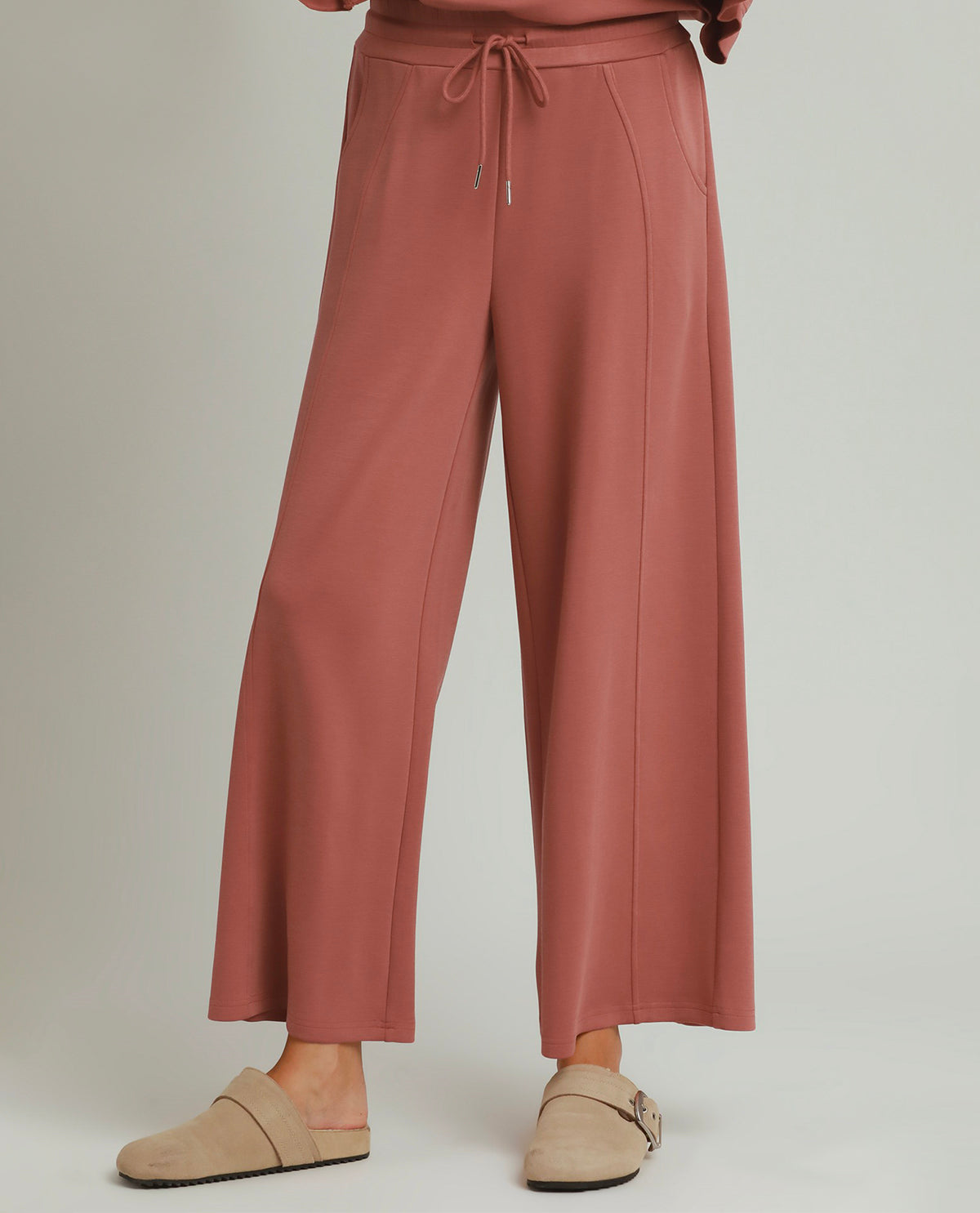 A person wearing dusty rose wide-leg drawstring pants and beige slip-on mule shoes, shown from the waist down against a light gray studio background.