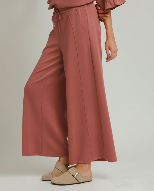 A model wearing dusty rose wide-leg cropped pants with a drawstring waist, paired with beige mule loafers, shown from the waist down on a light gray studio background.