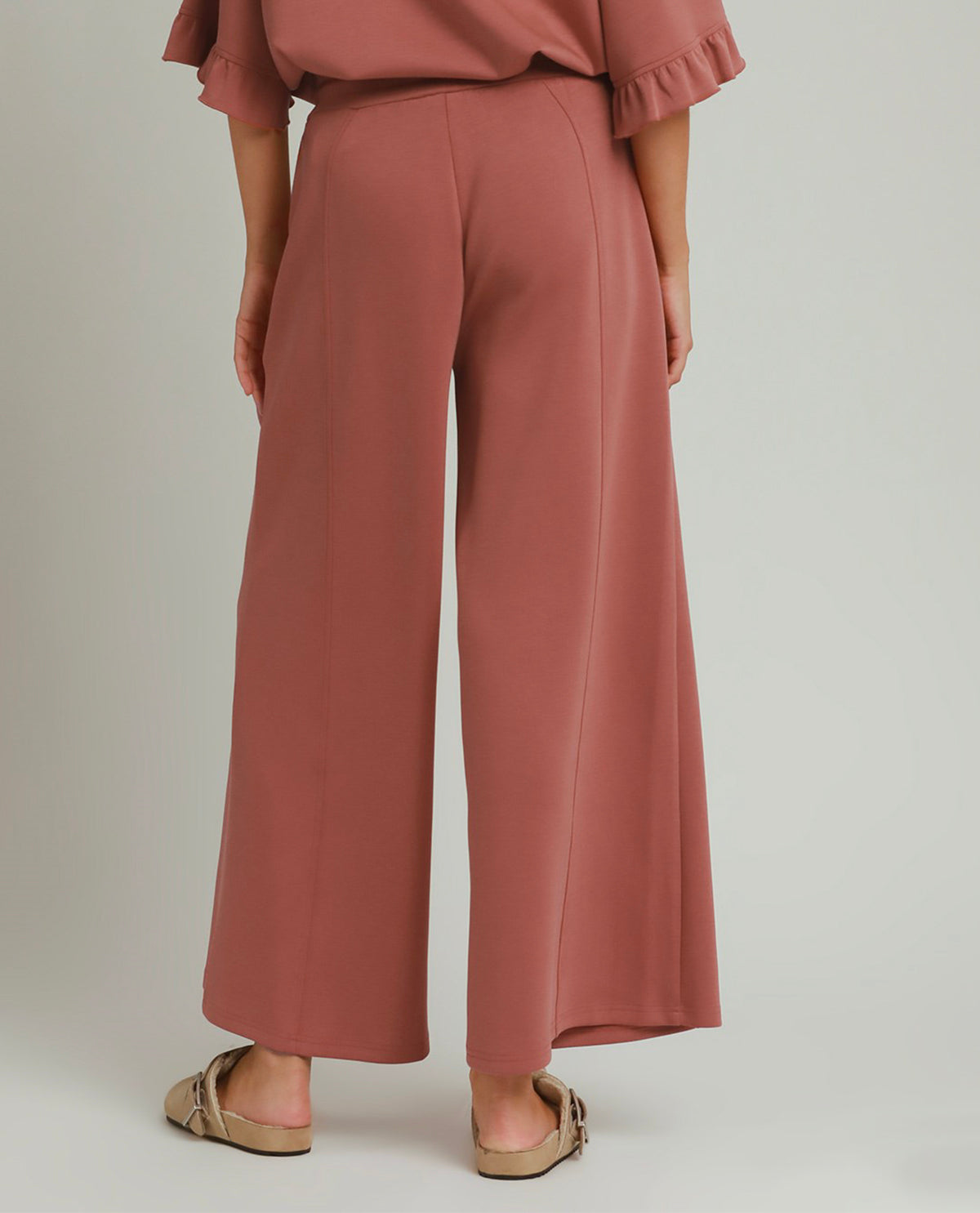 A person shown from the back wearing dusty rose wide-leg cropped pants with a matching ruffled blouse and beige flat sandals, photographed against a plain light gray studio background.