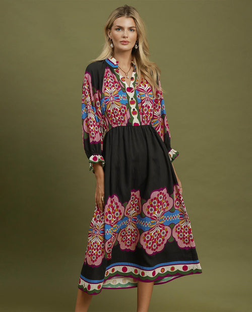 A woman modeling a long-sleeved midi dress with a black base and vibrant pink, red, blue, and green floral patterns, set against a solid olive green studio background.