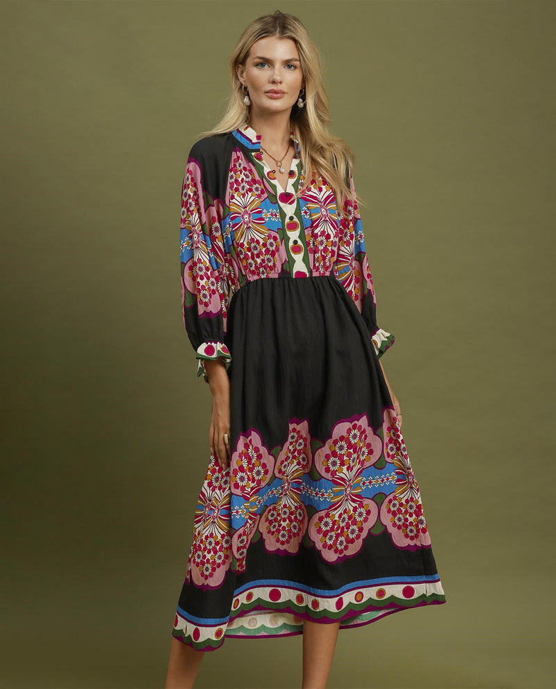 A woman modeling a long-sleeved midi dress with a black base and vibrant pink, red, blue, and green floral patterns, set against a solid olive green studio background.