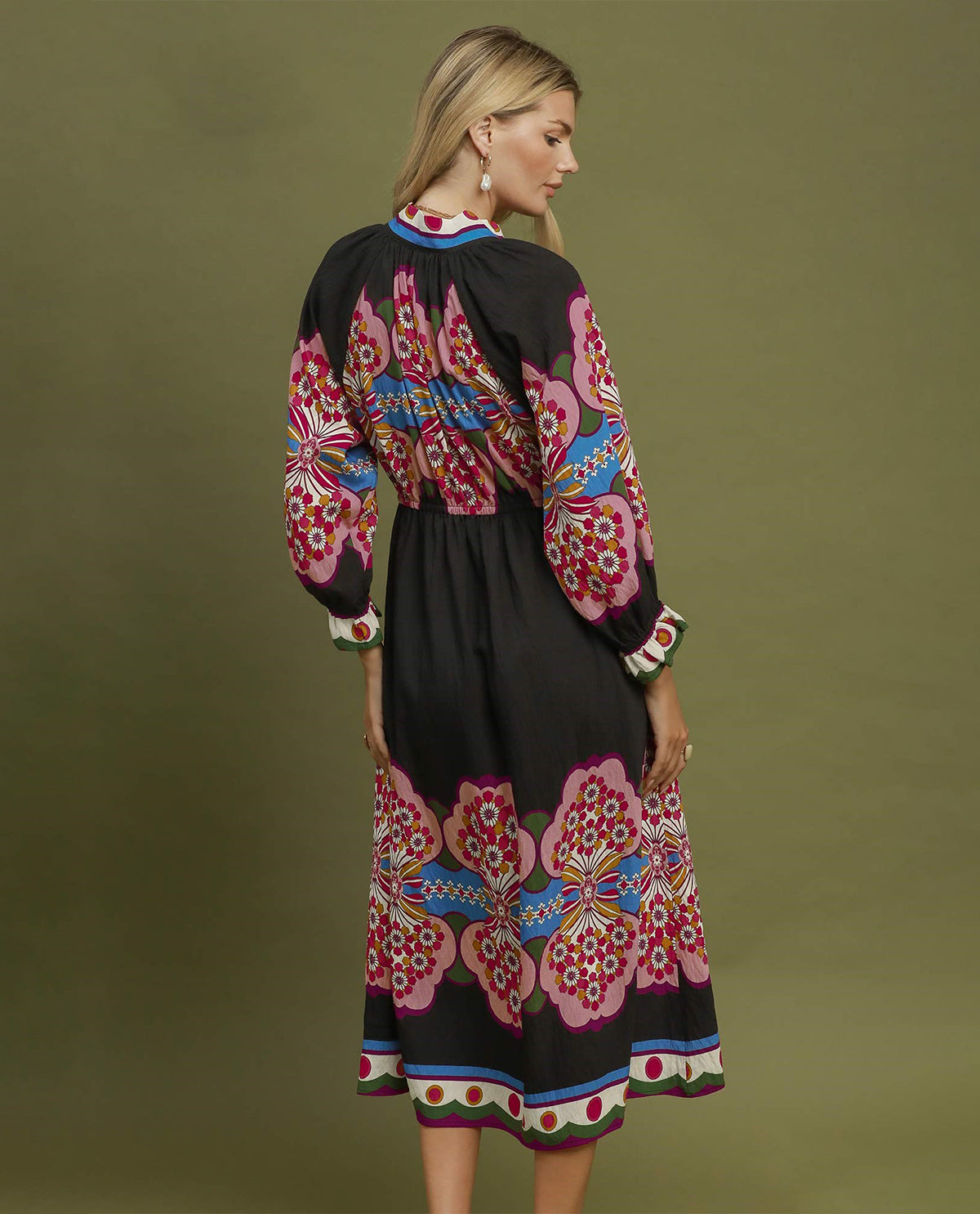 A woman modeling a black midi dress with vibrant pink, red, blue, and green floral embroidery, shown from a three-quarter back view against a solid olive green studio background.