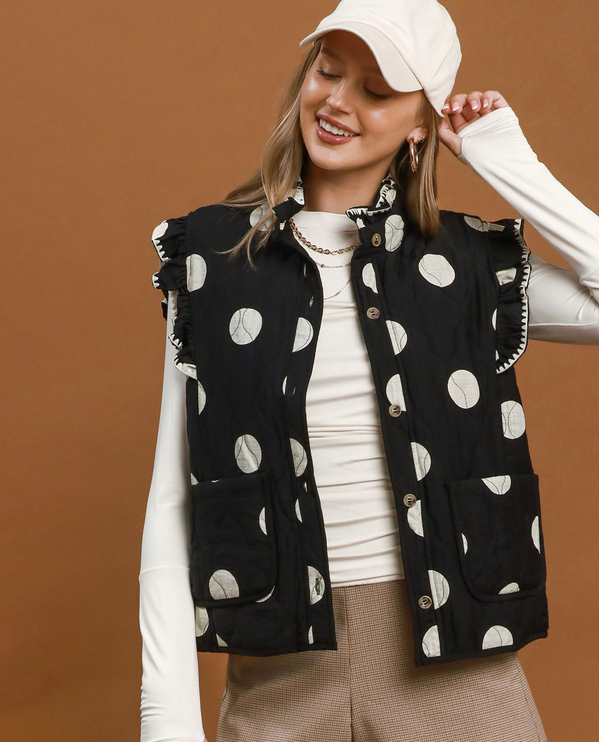 A woman wearing a black sleeveless vest with white moon-phase pattern, styled over a white long-sleeve top and beige pants against a solid brown studio background.