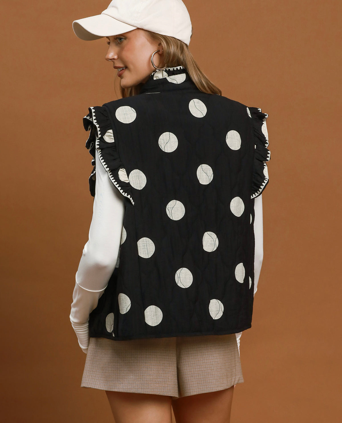 A woman shown from the back wearing a black sleeveless quilted vest with large white polka dots and ruffled trim, over a white long-sleeve top and beige shorts, against a solid brown studio background.