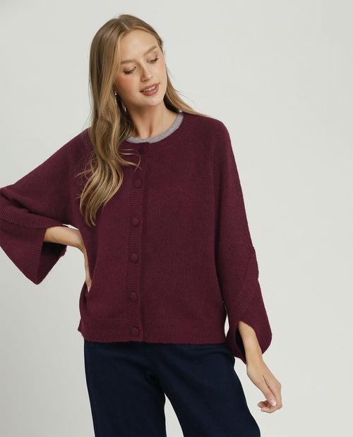 A woman wearing a burgundy knit cardigan with wide bell sleeves and a gray crew neck, paired with dark navy pants, against a light gray studio background.