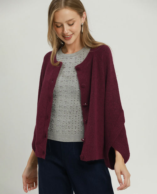 A woman wearing a burgundy knit cardigan over a gray knit top and navy pants, photographed against a plain light gray studio background.