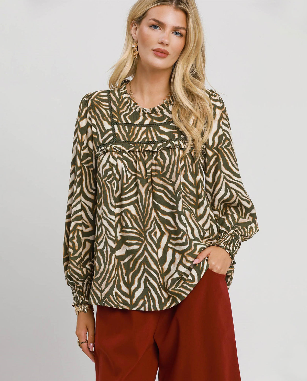 A woman modeling a long-sleeve blouse with an olive green, brown, and beige zebra-like pattern, paired with rust-colored pants, against a light gray studio background.