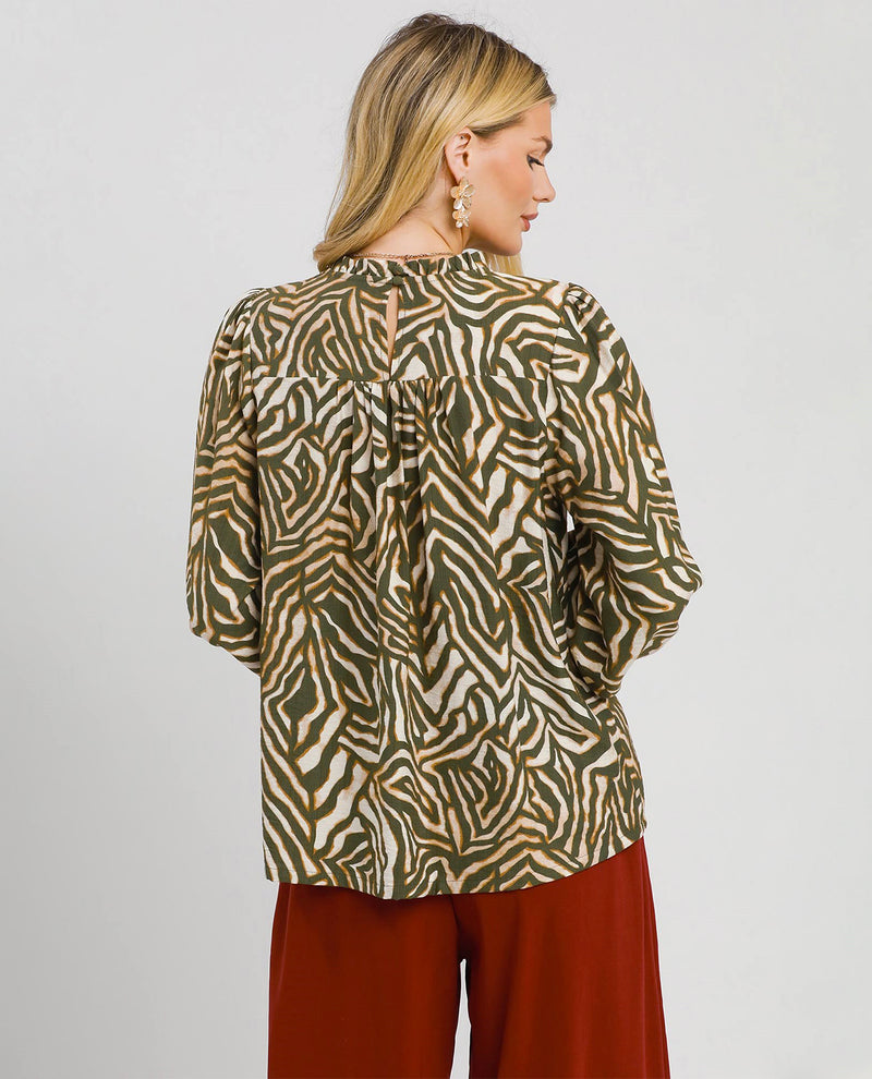A woman shown from the back wearing a zebra-print blouse with olive green, black, and beige stripes, paired with rust-red pants, against a light gray studio background.