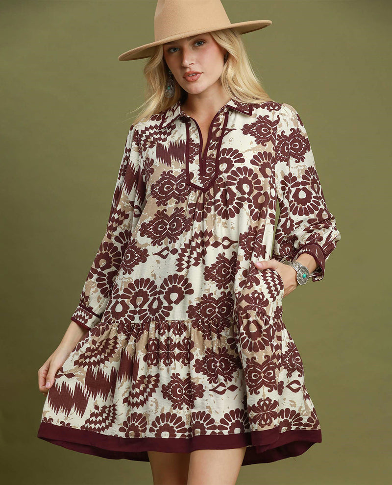 A woman modeling a bohemian-style mini dress with a brown and cream floral ikat pattern, wearing a tan wide-brim hat against a solid olive green background.