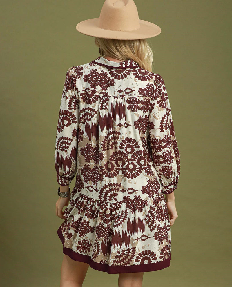 A woman viewed from behind wearing a long-sleeve bohemian-style dress with a cream and burgundy ikat pattern, paired with a beige wide-brim hat, against a solid olive green background.