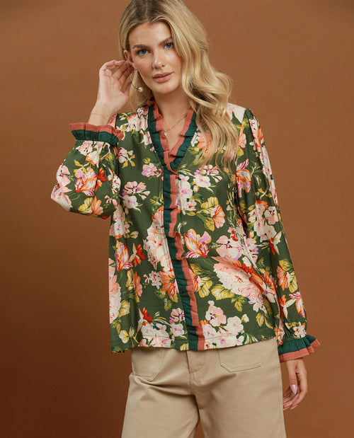 A woman models a long-sleeve button-up blouse with a green base and a vibrant floral print in pink, orange, and white, featuring ruffle trim. She is paired with beige trousers against a solid brown studio background.