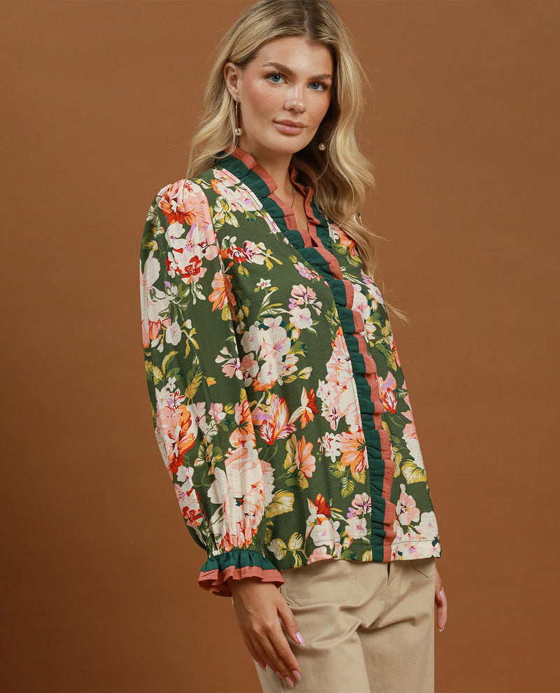 A woman modeling a long-sleeve button-up blouse with a green base and a vibrant multicolor floral print, paired with beige trousers, against a solid brown studio background.