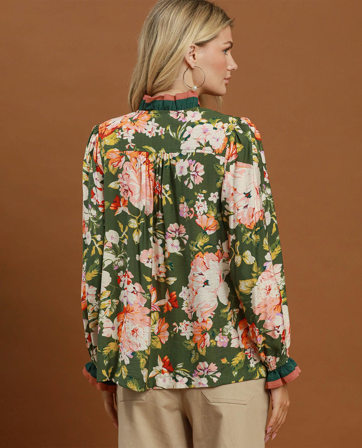 A woman shown from the back wearing a dark green long-sleeve blouse with a vibrant floral print in pink, orange, and white, paired with beige trousers, set against a solid brown studio background.
