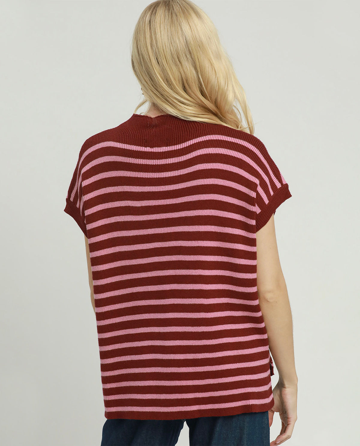 A woman viewed from behind wearing a short-sleeve knit top with horizontal burgundy and pink stripes, paired with blue jeans, against a plain light gray studio background.