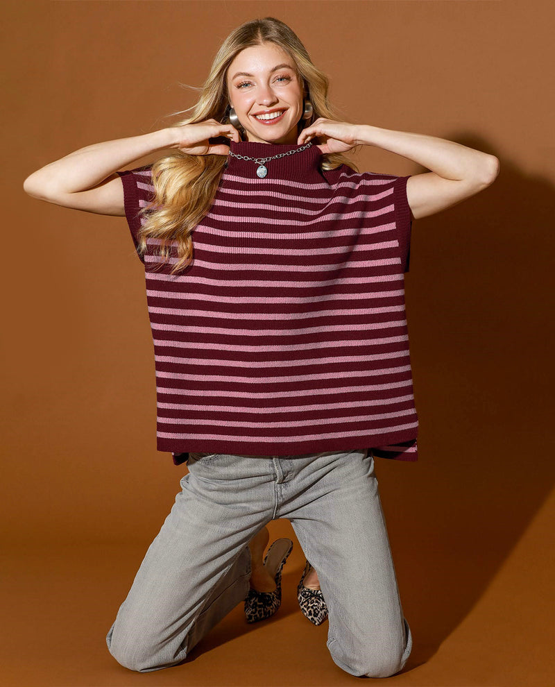 A woman kneeling against a solid brown studio background, wearing a sleeveless knit top with horizontal burgundy and dusty rose stripes, light gray jeans, and leopard-print heels.
