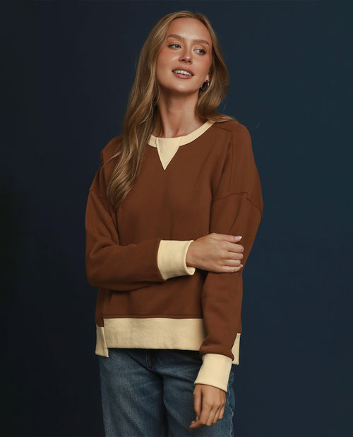 A woman modeling a brown long-sleeve knit sweater with contrasting cream panels at the V-neck, cuffs, and hem, paired with blue jeans against a dark navy studio background.