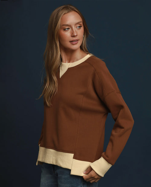 A woman modeling a brown long-sleeve sweater with contrasting cream-colored collar, hem, and cuffs, paired with blue jeans against a dark navy studio background.