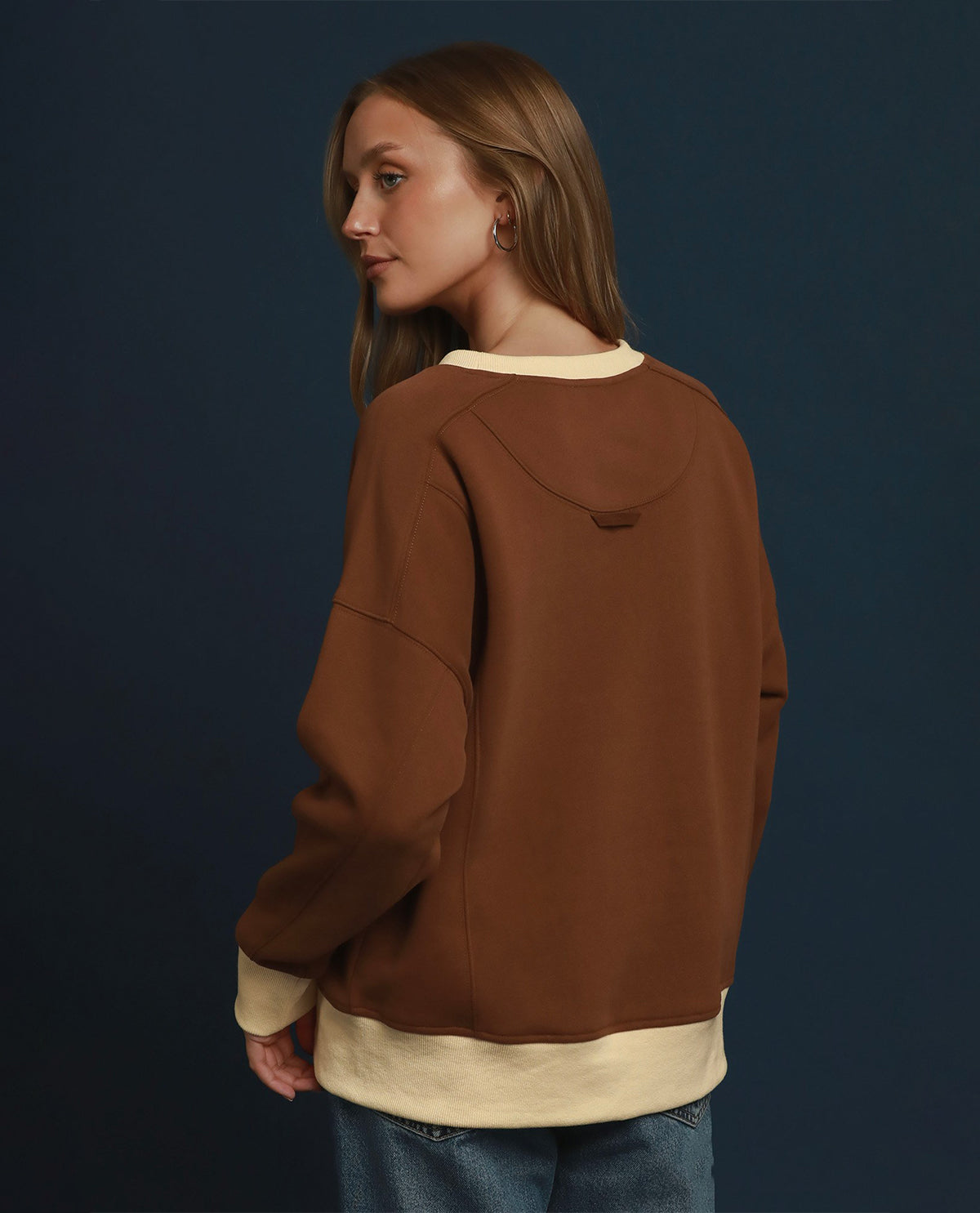 A woman wearing a brown oversized sweatshirt with cream ribbed trim, shown from the back against a dark blue studio background.