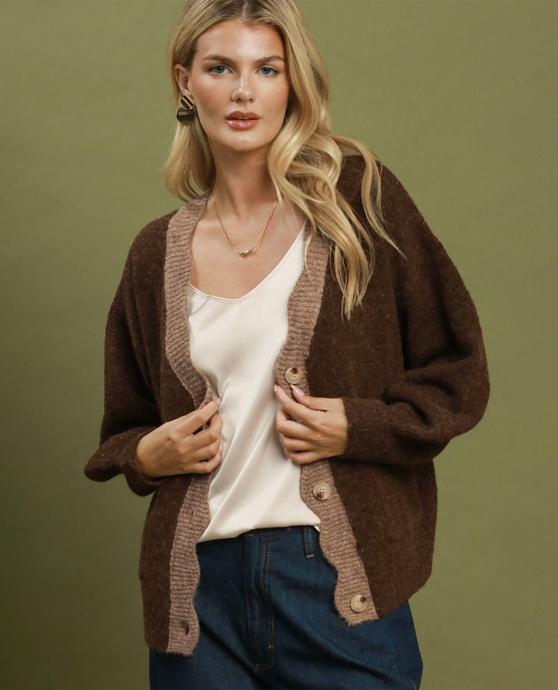 A woman models a brown open-front cardigan with ribbed trim over a cream camisole and blue jeans, set against a solid olive green studio background.