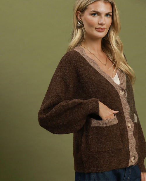 A woman shown from the side wearing a loose, oversized brown knit sweater with a wide neckline, paired with dark blue jeans, against a solid olive green studio background with hands in pockets on cardigan.