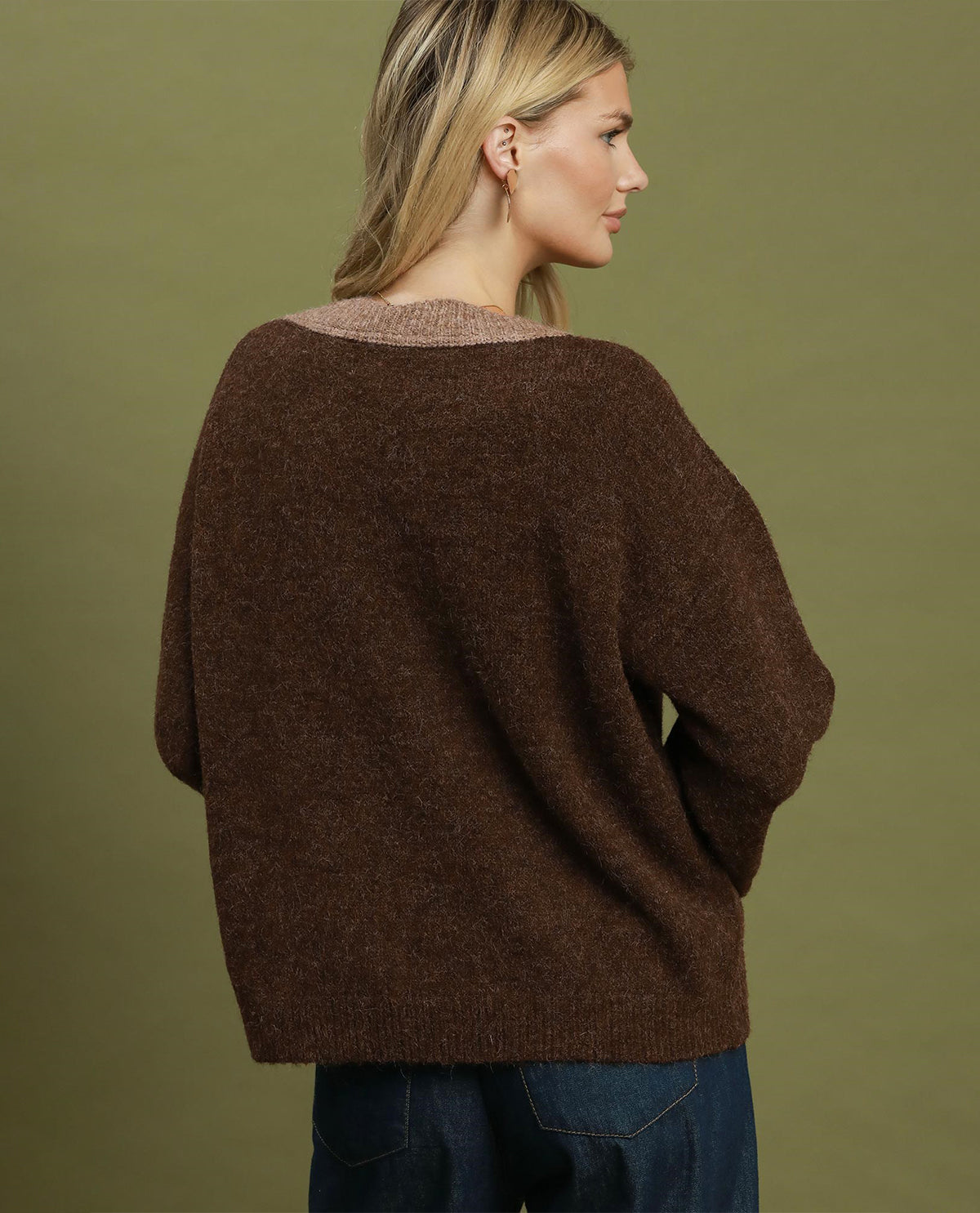 A woman shown from the back wearing a loose, oversized brown knit sweater with a wide neckline, paired with dark blue jeans, against a solid olive green studio background.