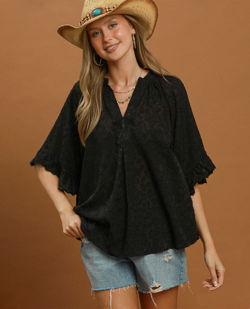 A woman wearing a black lace short-sleeve blouse with ruffled cuffs, light-wash denim shorts, and a straw cowboy hat, posed against a solid brown studio background.