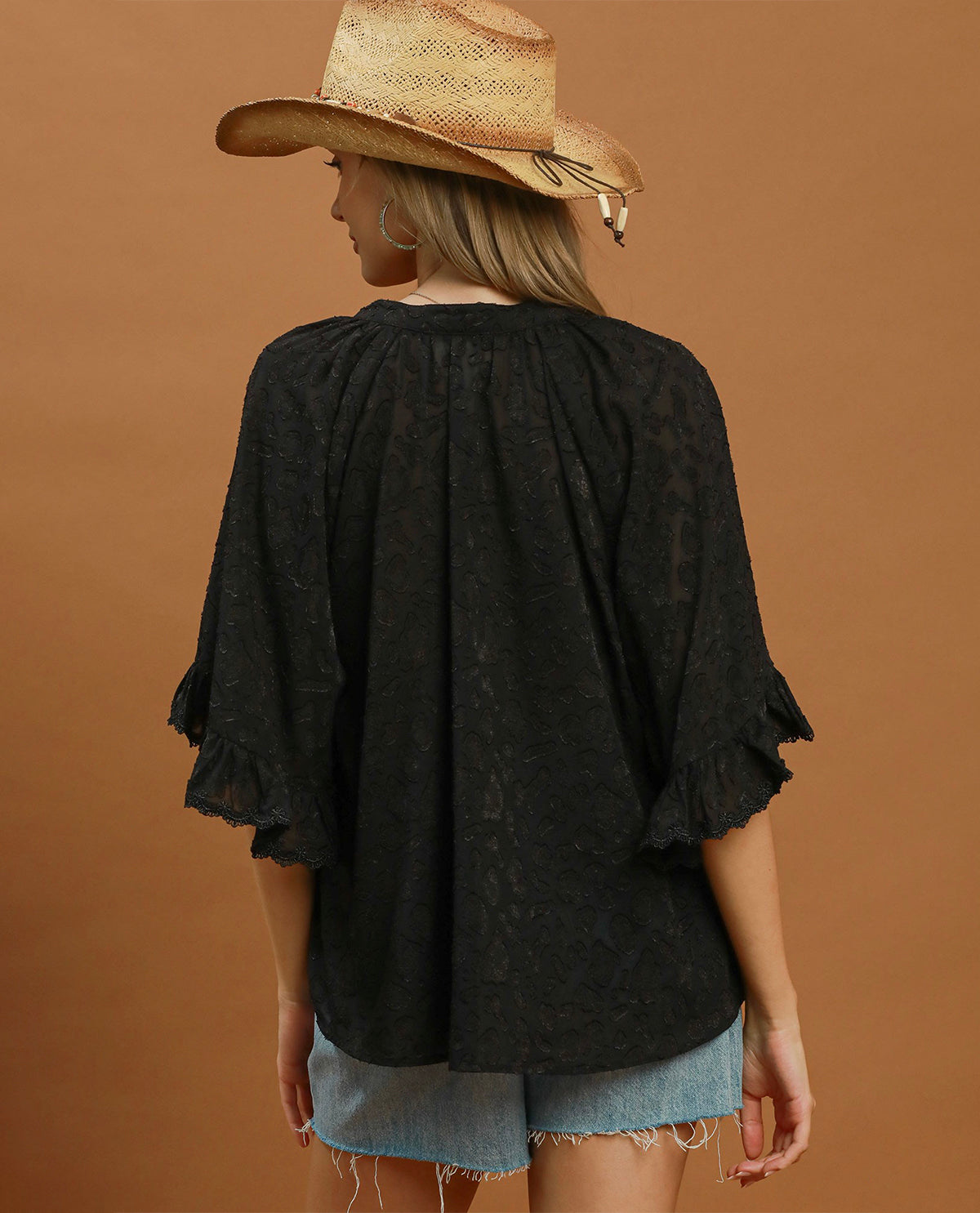 A woman shown from the back wearing a loose black lace blouse with ruffled elbow-length sleeves and light blue denim shorts, standing against a solid warm brown studio background.
