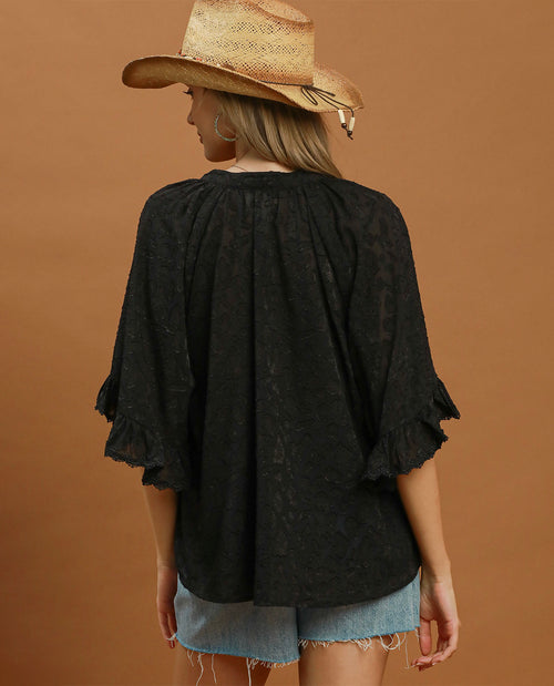 A woman shown from the back wearing a loose black lace blouse with ruffled elbow-length sleeves and light blue denim shorts, standing against a solid warm brown studio background.