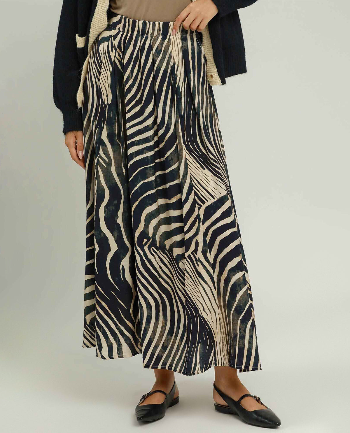 A model wearing a high-waisted, long maxi skirt with a black and beige zebra-like wavy pattern, styled with a beige top, a dark cardigan, and black flat shoes against a light gray studio background.