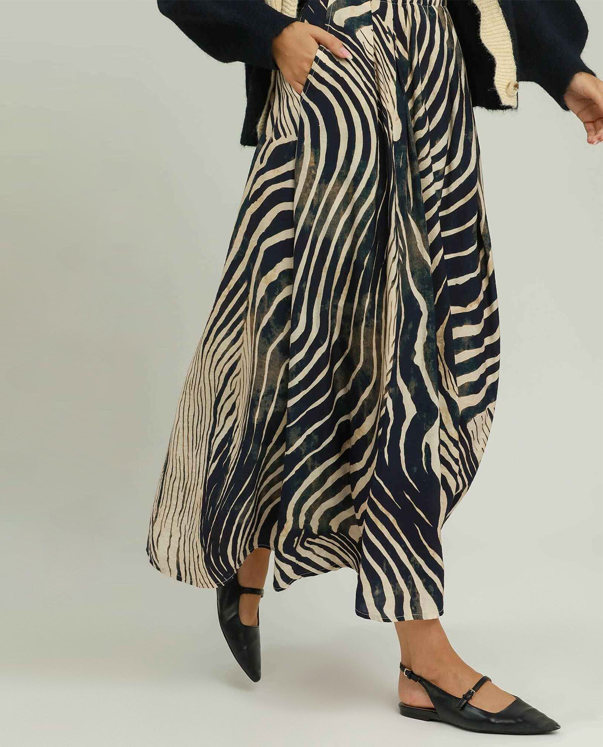 A person wearing a high-waisted, wide-leg maxi skirt with a bold black and beige zebra-like abstract pattern, styled with a dark cardigan and black flat shoes against a light gray studio background.