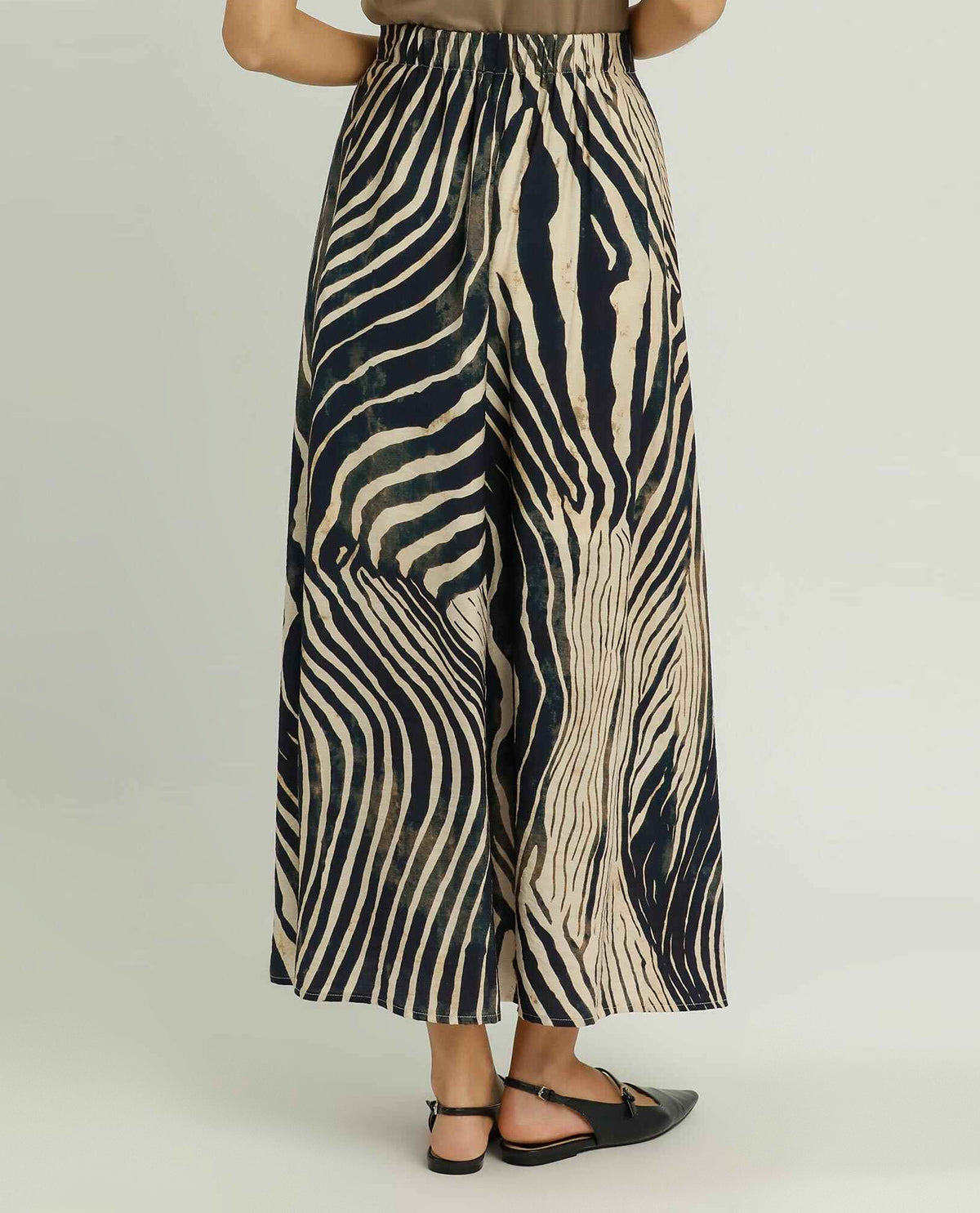 A person shown from the waist down wearing wide-leg cropped pants with a bold zebra-like abstract stripe pattern in black and warm beige, paired with black flat sandals against a light gray studio background.