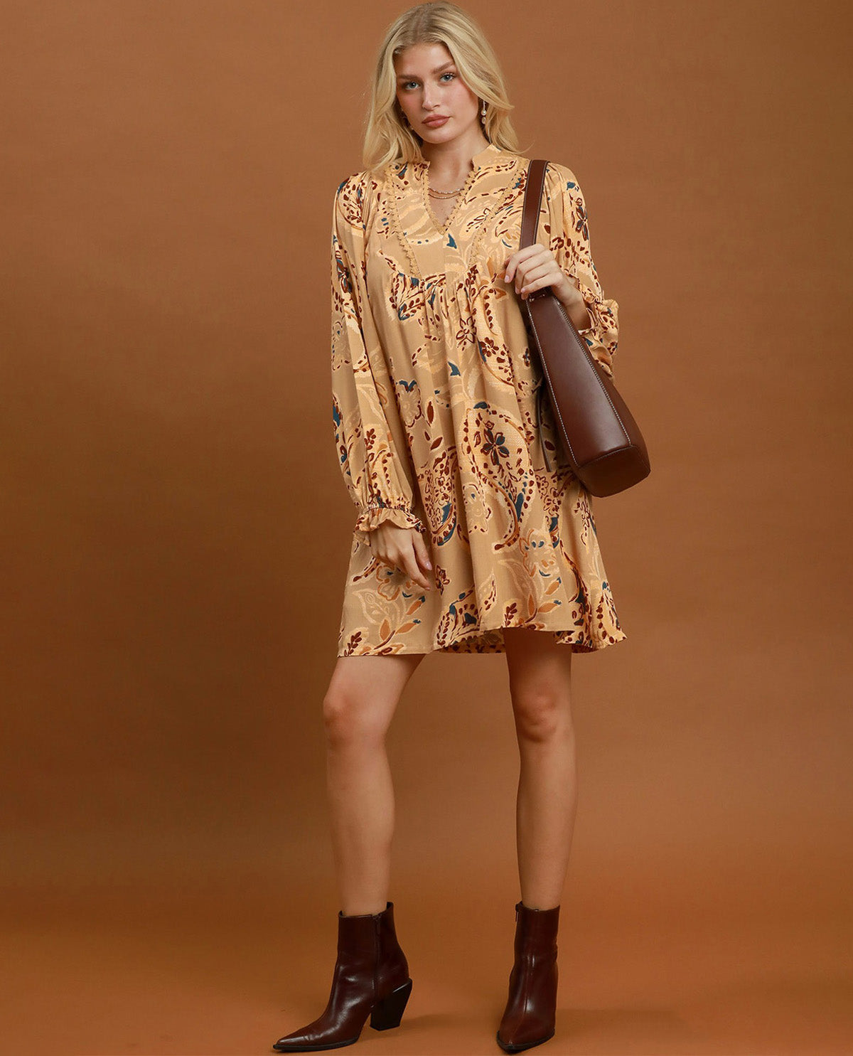 A woman modeling a beige long-sleeve mini dress with a brown and black paisley pattern, holding a brown leather shoulder bag and wearing dark brown ankle boots, set against a solid brown studio background.
