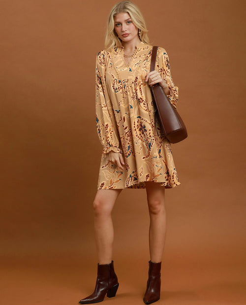A woman modeling a beige long-sleeve mini dress with a brown and black paisley pattern, holding a brown leather shoulder bag and wearing dark brown ankle boots, set against a solid brown studio background.