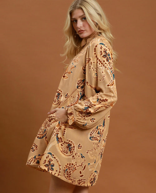 A woman modeling a long-sleeve beige tunic dress with a brown and blue paisley pattern, posed against a solid warm brown studio background.