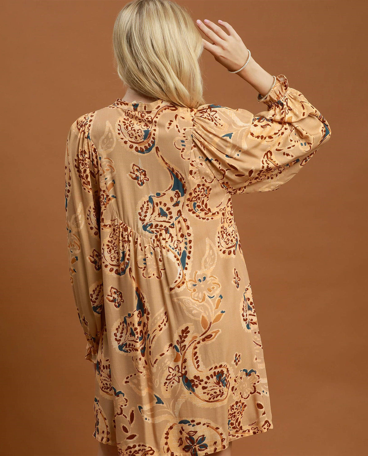 A woman shown from the back wearing a beige long-sleeve tunic dress with a brown and blue paisley pattern, set against a solid brown studio background.