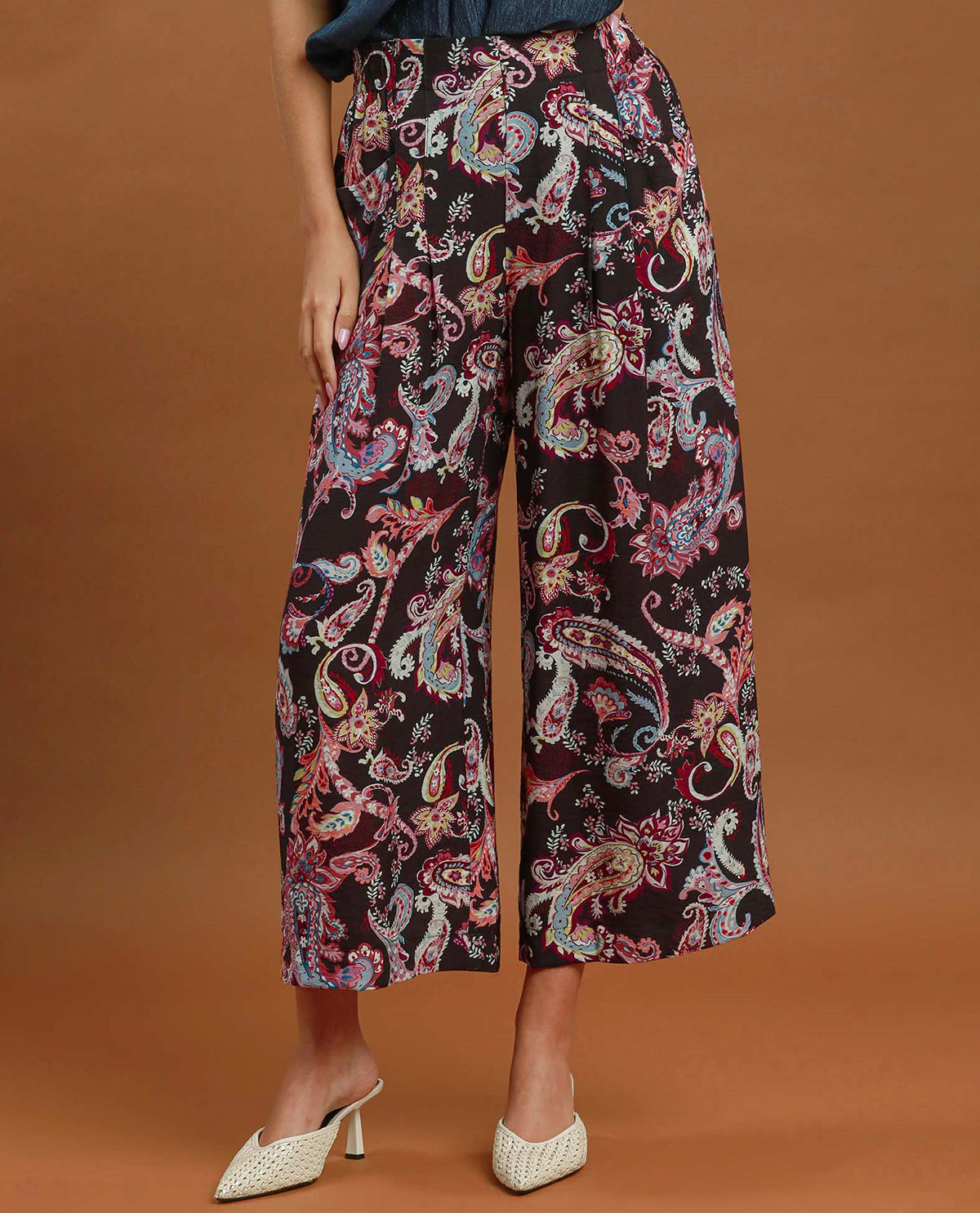 A model is shown from the waist down wearing high-waisted, wide-leg cropped pants with a vibrant paisley pattern on a black base, paired with white textured mule heels against a solid brown studio background.